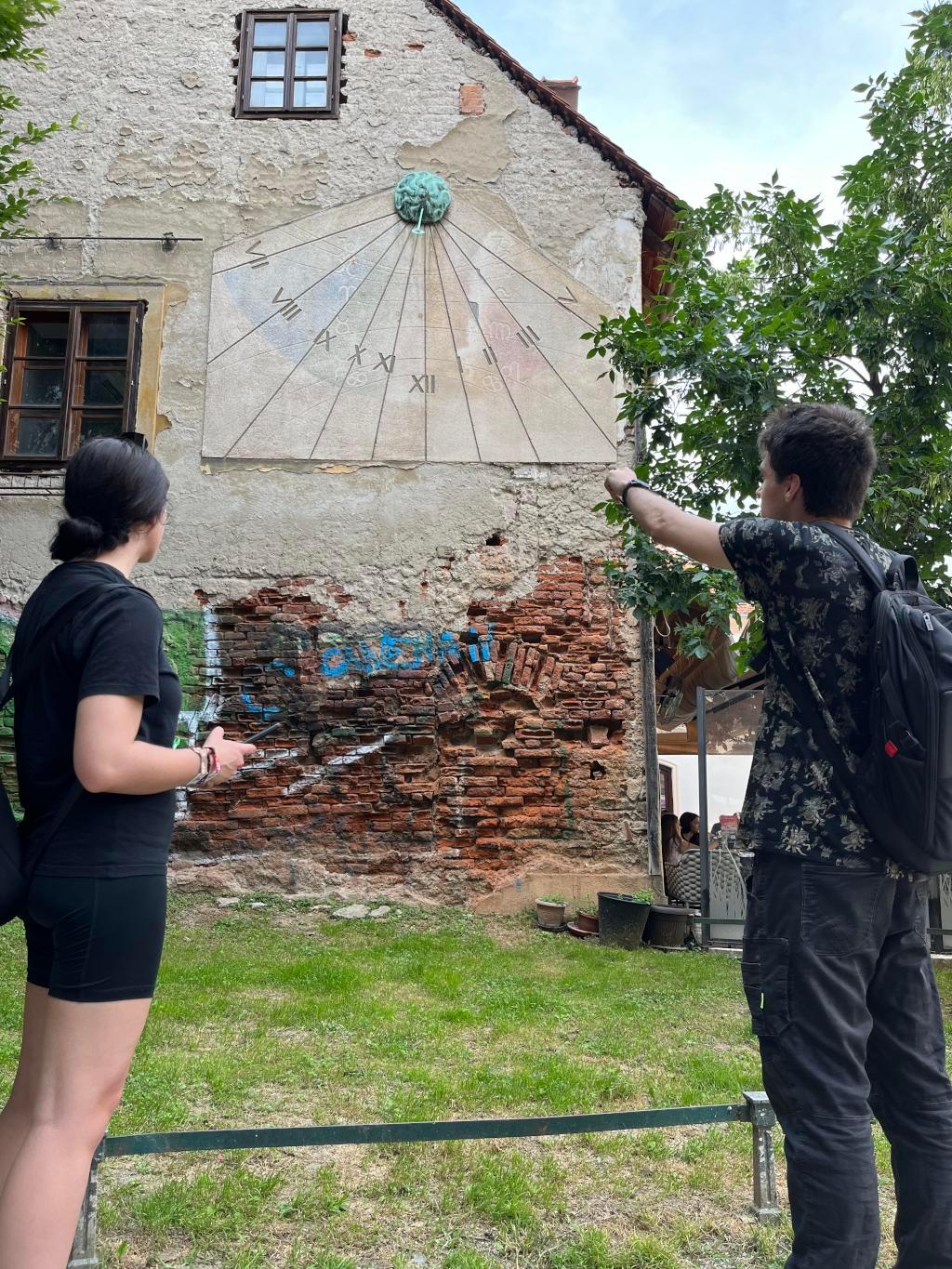 A man pointing at a sun clock and woman looking for clues.